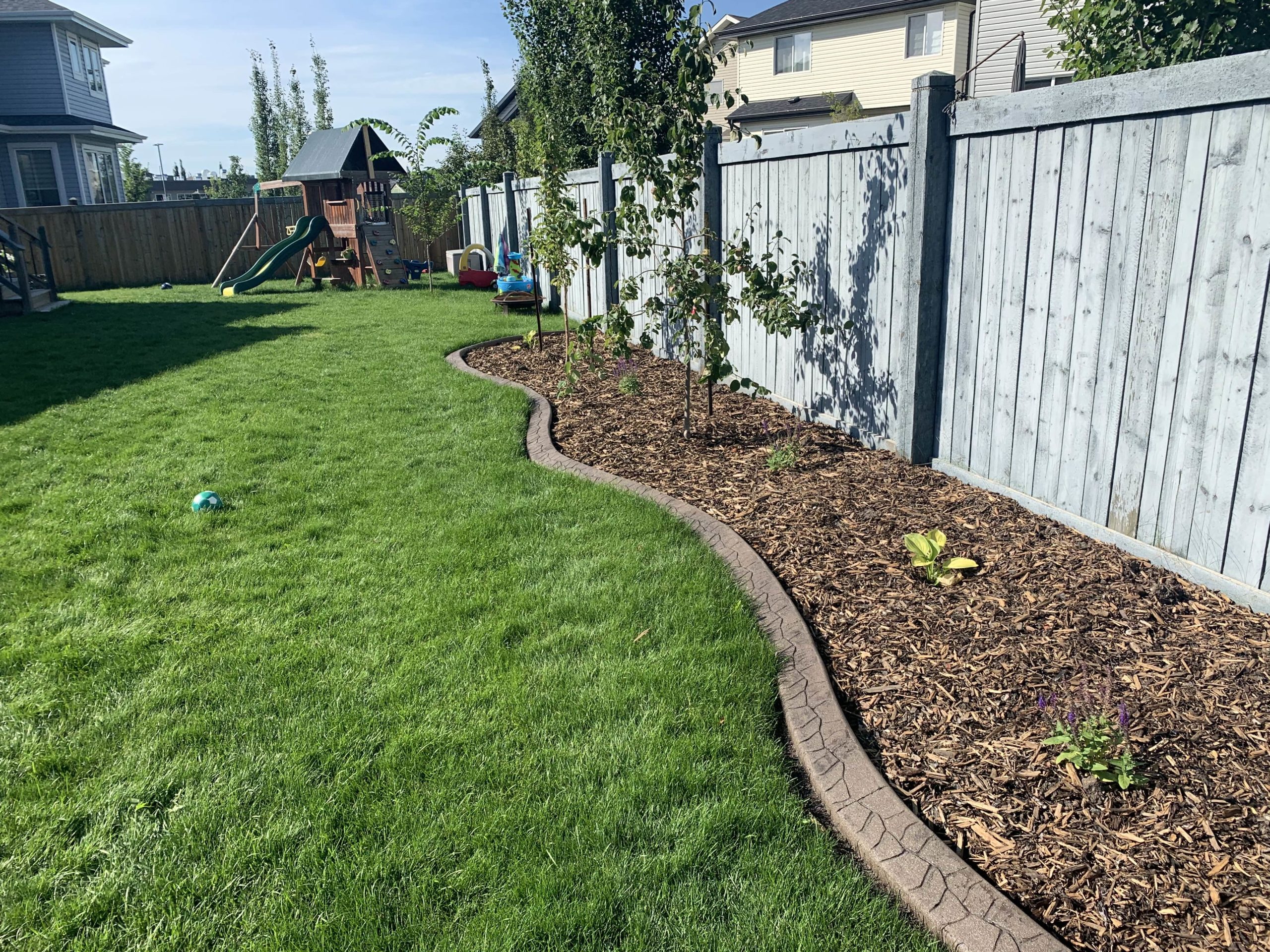 backyard-landscaping-edmonton Landscaping construction in a backyard with very green grass and a mulch bed along the fence. Concrete curbing is along the mulch bed.